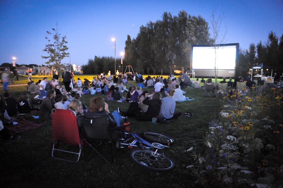 People sitting on grass watching a film outdoors at dusk, bicycles and flowers nearby.